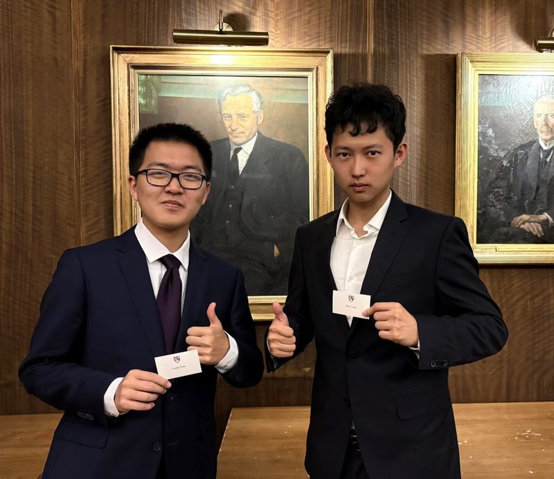 Two students in suits holding their cards in a panelled room
